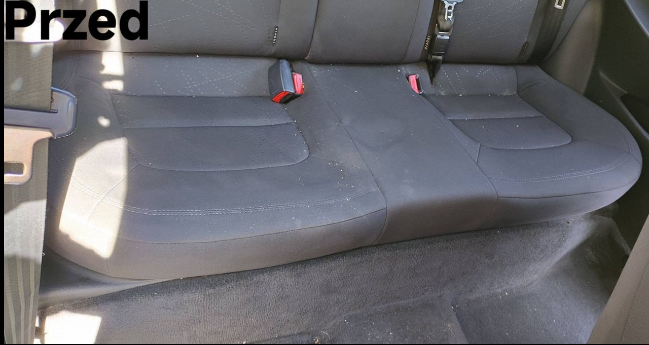 Zabrudzona tylna kanapa samochodu przed czyszczeniem. Widoczne okruchy i plamy na ciemnej tapicerce. Ujęcie z bliska, z napisem 'Przed' w lewym górnym rogu.