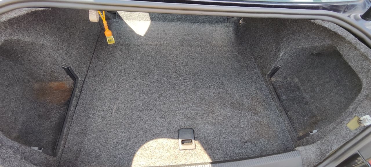 Wnętrze bagażnika samochodu wyłożone szarą wykładziną dywanową. Widoczne elementy mocujące i uchwyt. Jasne plamy światła podkreślają strukturę materiału.