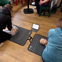NAST Sp. z o.o. - Kurs pierwszej pomocy: uczestnicy ćwiczą resuscytację na fantomach pod okiem instruktora. Tablet z instrukcjami leży obok. Jasne wnętrze, drewniana podłoga.