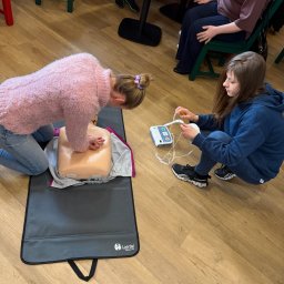 NAST Sp. z o.o. - Kurs pierwszej pomocy: Kobieta ćwiczy resuscytację na manekinie, druga przygotowuje defibrylator. Szkolenie z udzielania pierwszej pomocy w jasnym pomieszczeniu.