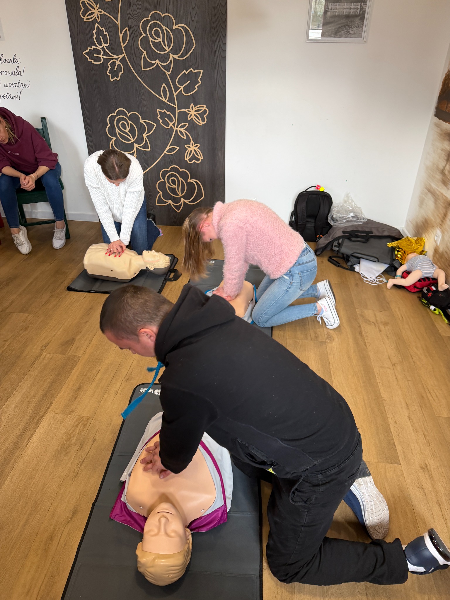 Kurs pierwszej pomocy: grupa osób ćwiczy resuscytację krążeniowo-oddechową na manekinach w jasnym pomieszczeniu z drewnianą podłogą i dekoracyjną ścianą.