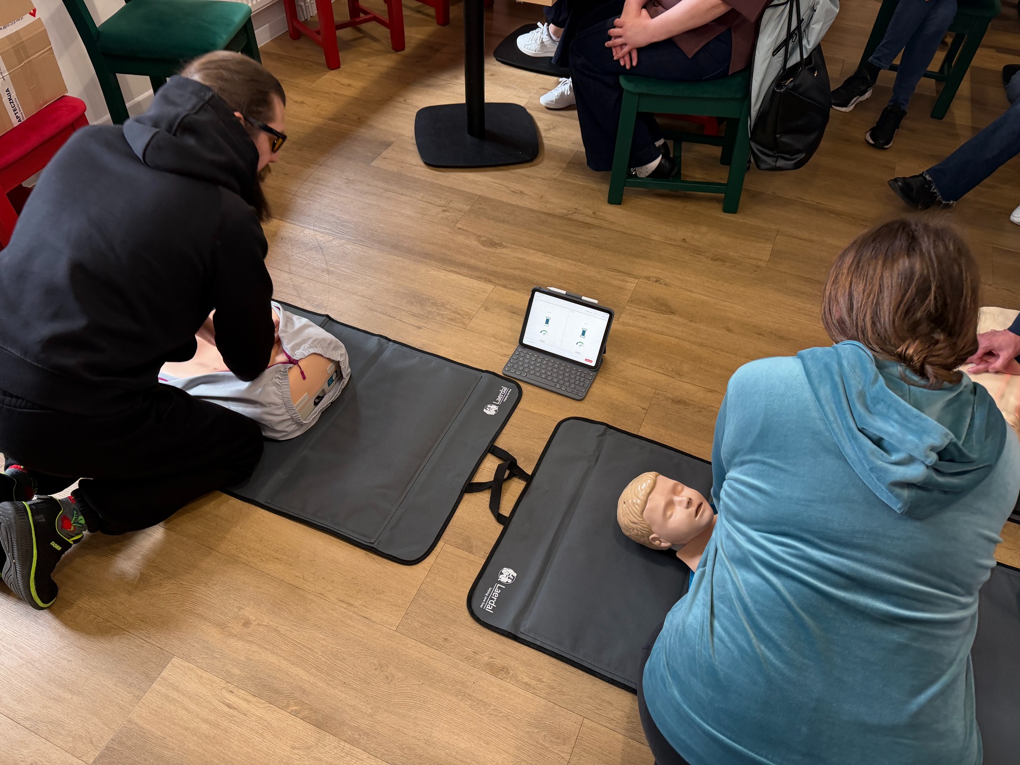 Kurs pierwszej pomocy: uczestnicy ćwiczą resuscytację na fantomach pod okiem instruktora. Tablet z instrukcjami leży obok. Jasne wnętrze, drewniana podłoga.