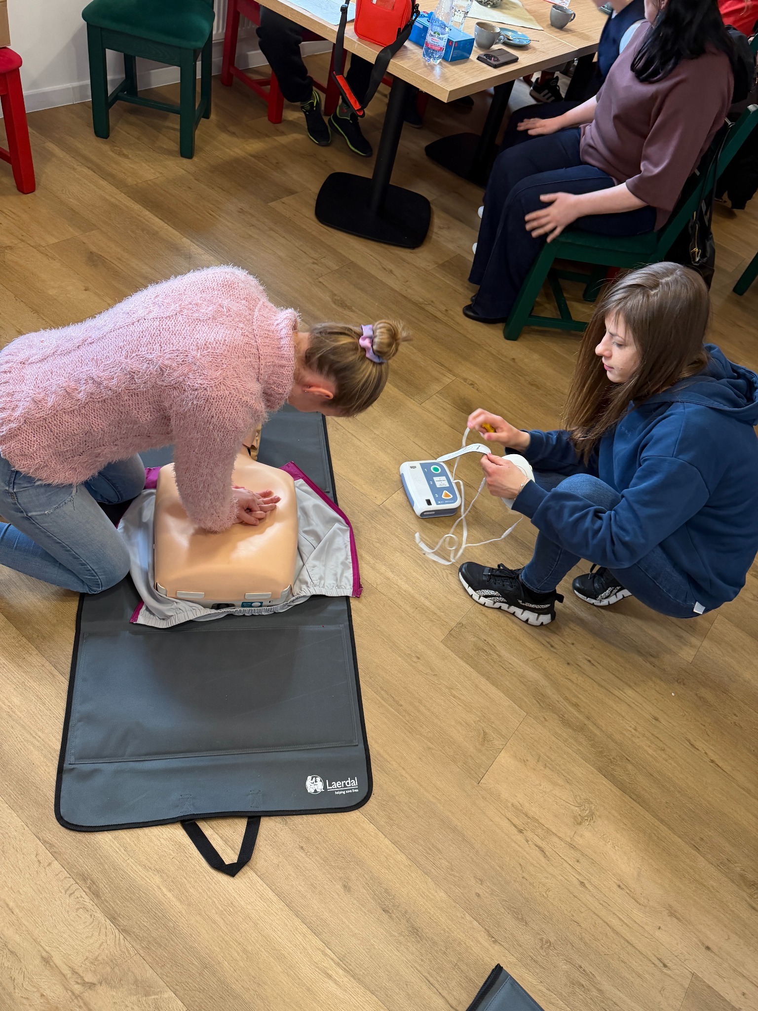 Kurs pierwszej pomocy: Kobieta ćwiczy resuscytację na manekinie, druga przygotowuje defibrylator. Szkolenie z udzielania pierwszej pomocy w jasnym pomieszczeniu.