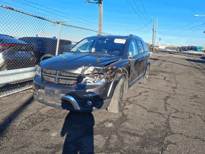 Uszkodzony, czarny Dodge Journey z widocznymi wgnieceniami i zarysowaniami na przednim błotniku i masce, stojący na parkingu w słoneczny dzień.