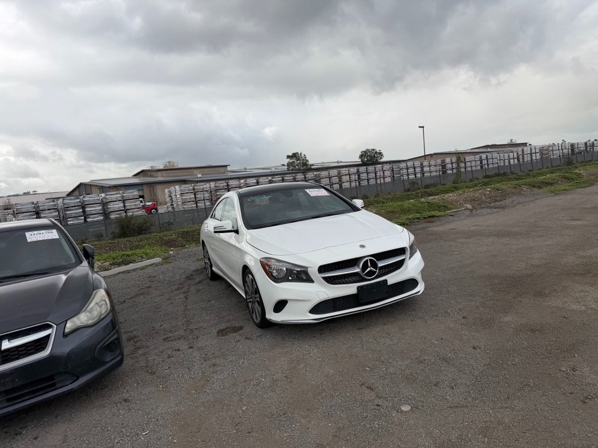 Biały Mercedes CLA na żwirowym parkingu, obok szary Subaru. W tle budynek magazynowy i pochmurne niebo. Widoczne naklejki na szybach aut.