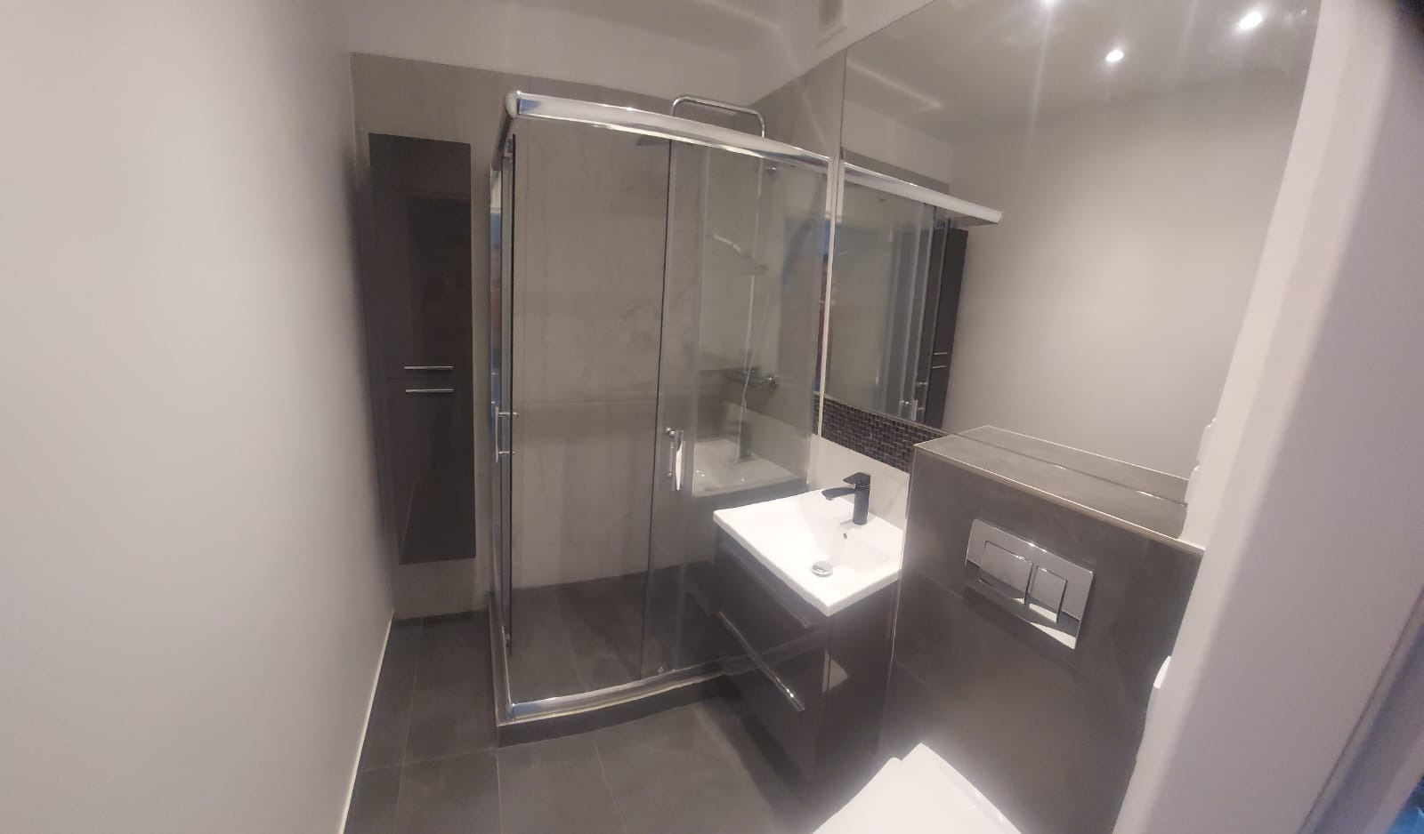 Nowoczesna łazienka po remoncie: kabina prysznicowa ze szklanymi drzwiami, umywalka z szafką, lustro z mozaiką i szara kolorystyka ścian i podłogi.