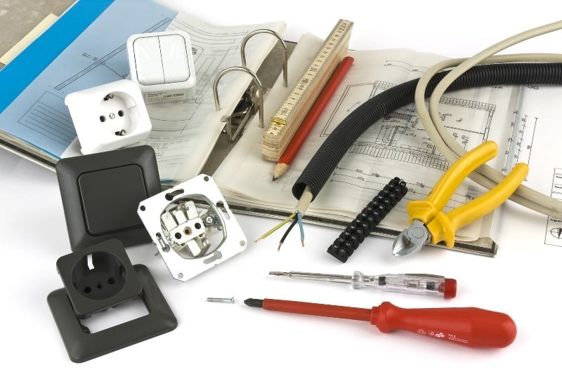 Elementy instalacji elektrycznej: gniazdka, włączniki, przewody, narzędzia, złączki, miernik, schemat instalacji w segregatorze na białym tle.
