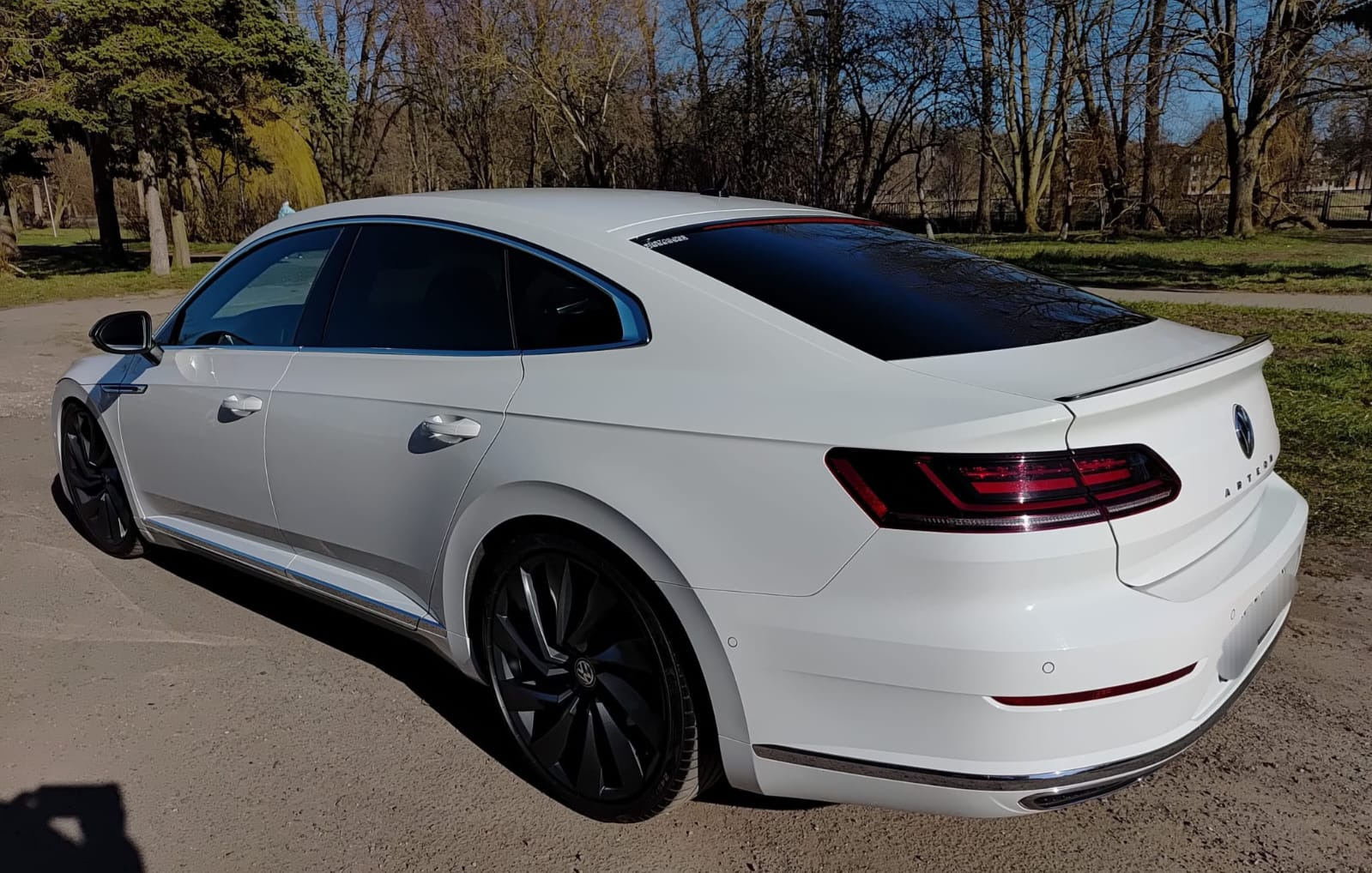 Biały Volkswagen Arteon po przyciemnieniu szyb, widoczny z tyłu pod kątem. Ciemne szyby kontrastują z jasnym lakierem. Tło stanowią drzewa i trawnik.
