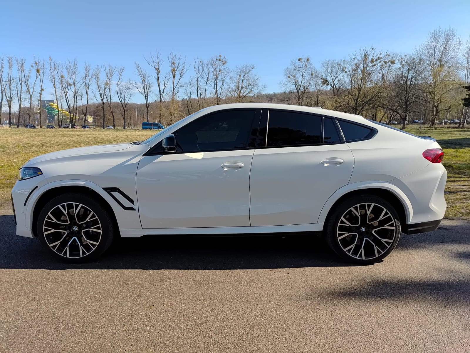 Białe BMW X6 z przyciemnionymi szybami bocznymi i tylną, zaparkowane na asfalcie w słoneczny dzień. Widok z boku, na tle drzew i błękitnego nieba.