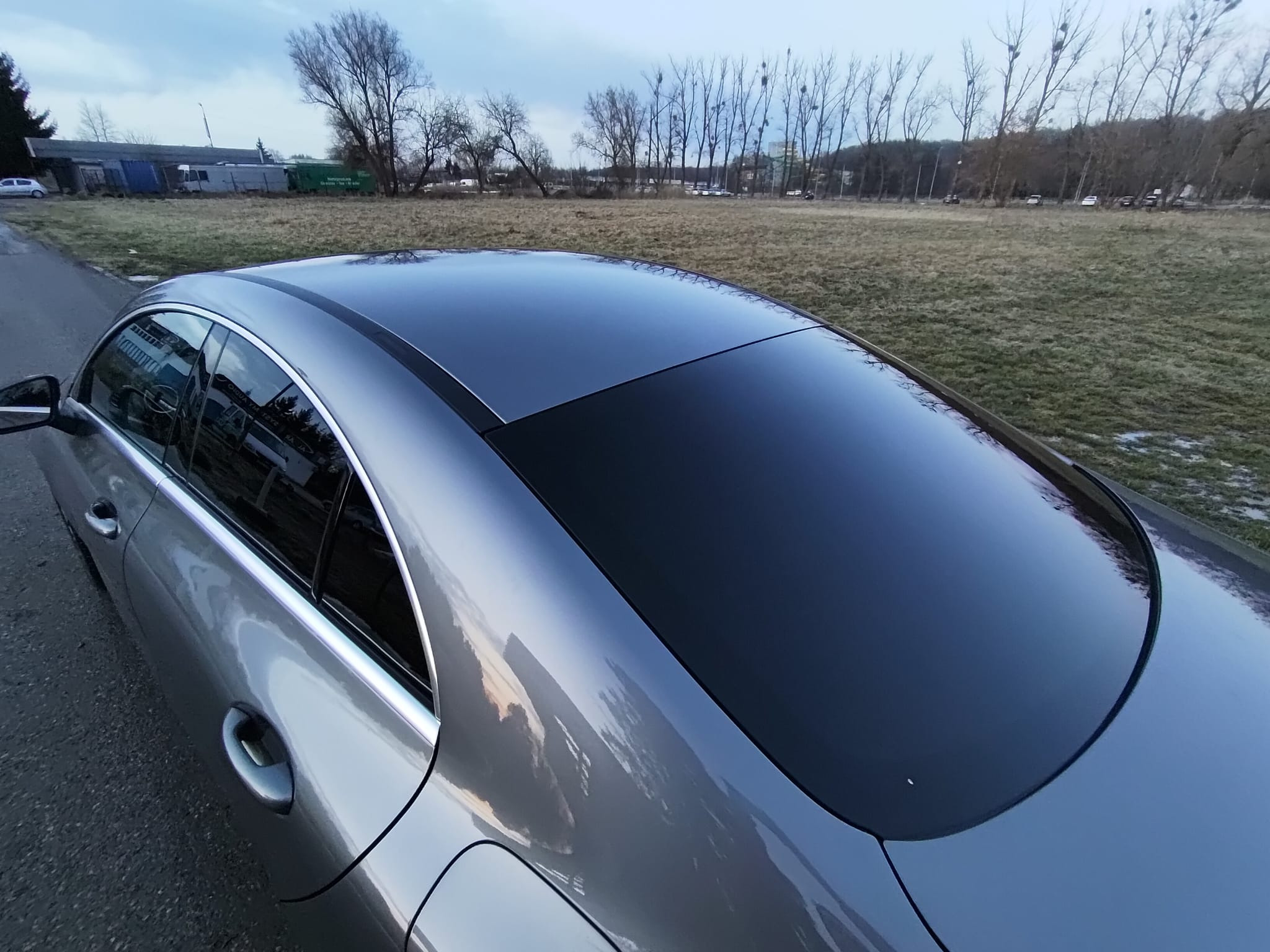 Szary samochód z przyciemnionymi szybami, widok z góry, na tle łąki i drzew. Szyby odbijają niebo, co podkreśla efekt.