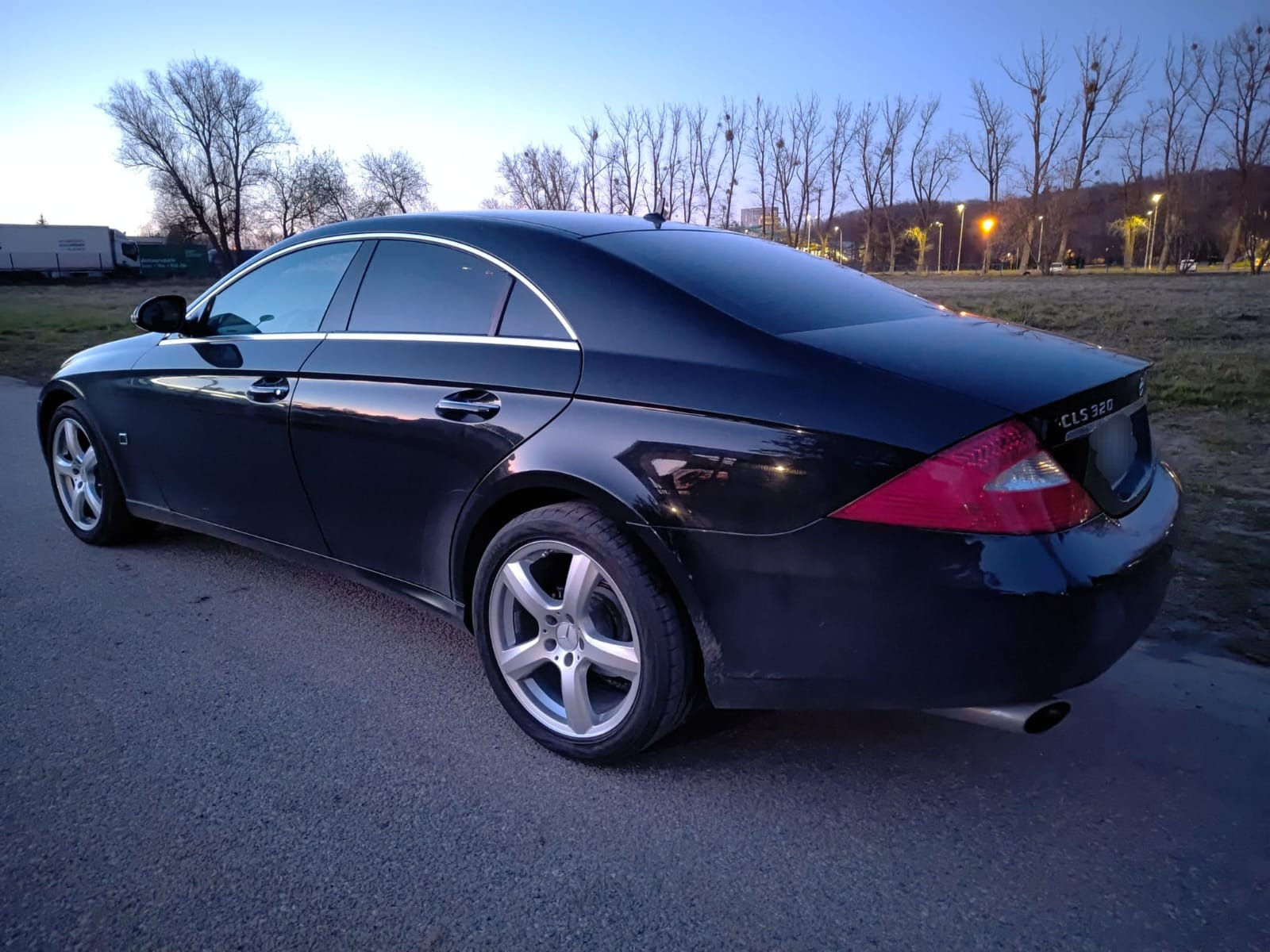 Czarny Mercedes CLS 320 po usłudze, widok z tyłu, lekko przyciemnione szyby, srebrne felgi, tło: drzewa i oświetlenie uliczne.