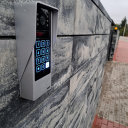 S-Tech Paweł Steć - Szary wideodomofon Eura z podświetlaną klawiaturą numeryczną zamontowany na szarym, teksturowanym murze z kostki, widok z boku, oświetlenie dzienne.
