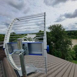 S-Tech Paweł Steć - Antena kierunkowa zamontowana na dachu domu z widokiem na rzekę i zieleń. Metalowa konstrukcja anteny z elementami białymi i niebieskimi. Pochmurne niebo w tle.
