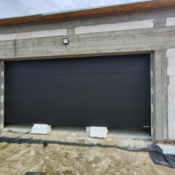 S-Tech Paweł Steć - Zamknięta, segmentowa brama garażowa w surowym otoczeniu budowy, z widocznym oświetleniem zewnętrznym i blokami podtrzymującymi. Szara kolorystyka bramy.