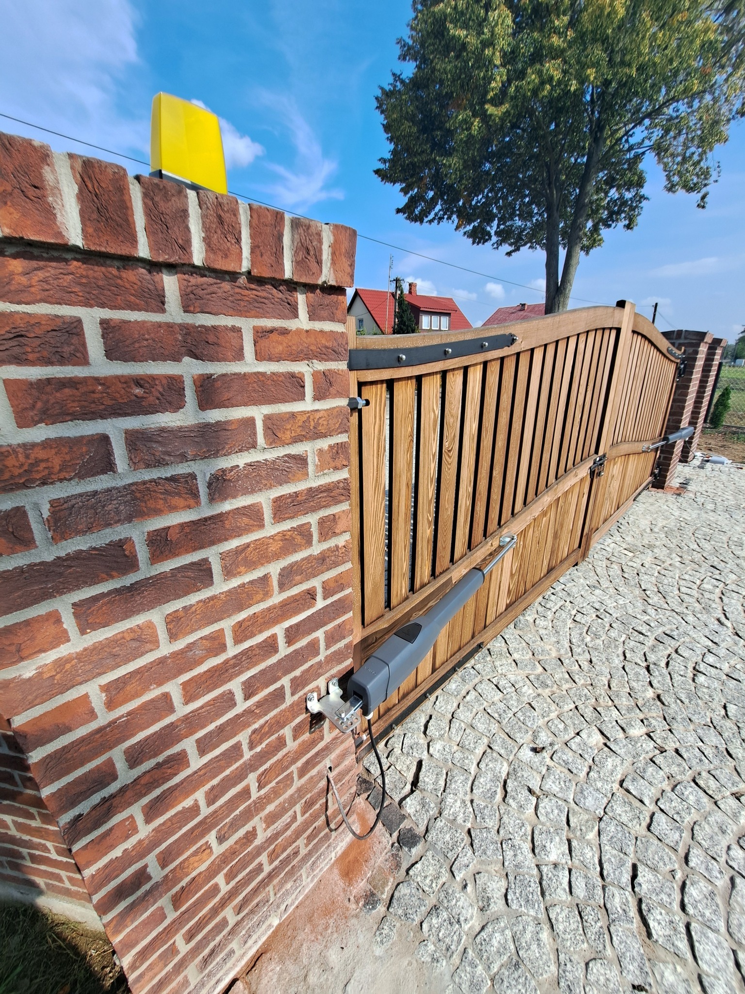 Drewniana brama wjazdowa z automatyką, zamontowana na ceglanym słupku. Widoczny siłownik bramy i lampa ostrzegawcza. Ułożona kostka brukowa.