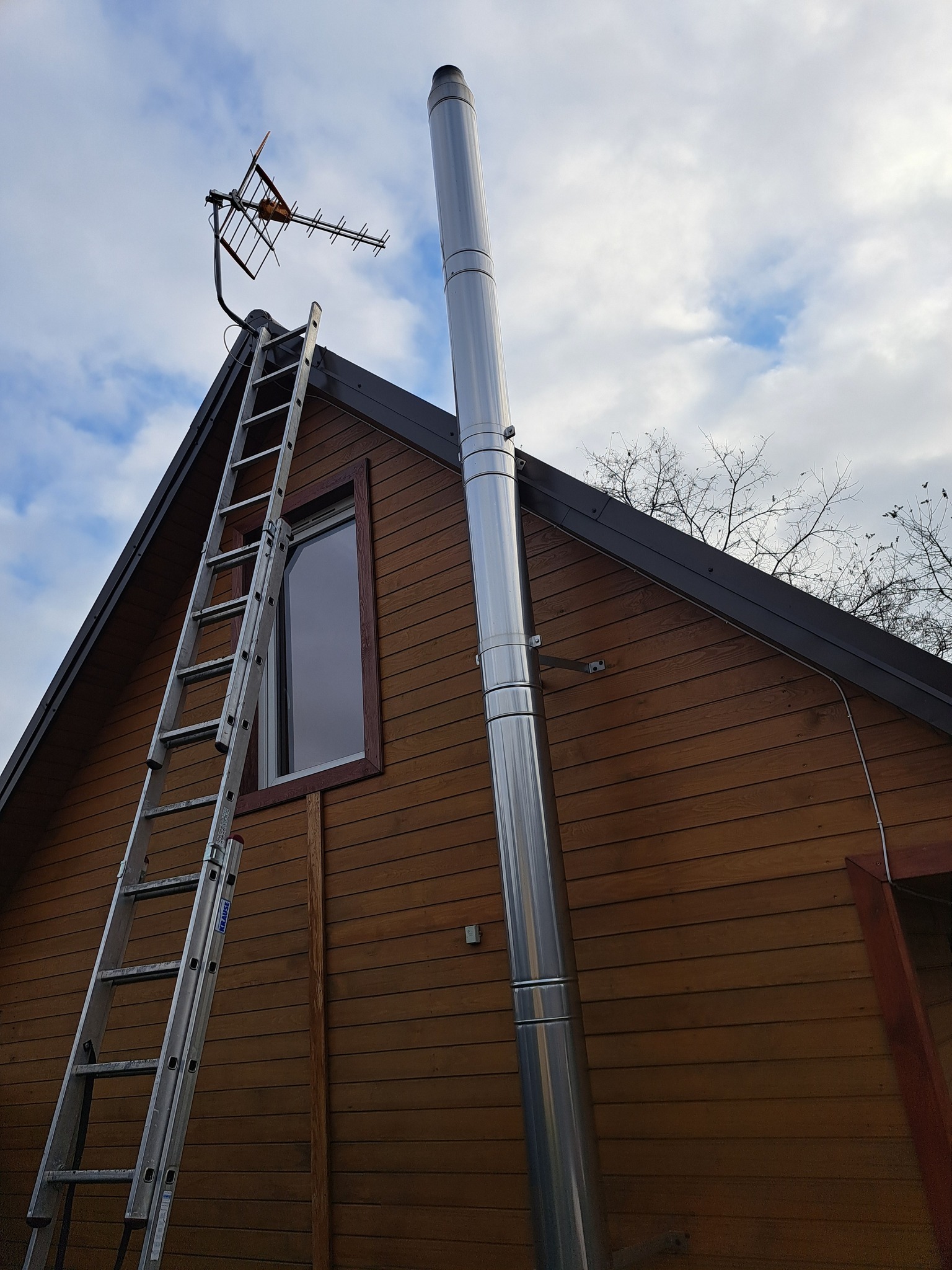 Antena telewizyjna na dachu drewnianego domu, wsparta drabiną. Widoczny komin ze stali nierdzewnej i fragment nieba z chmurami. Ujęcie z dołu.