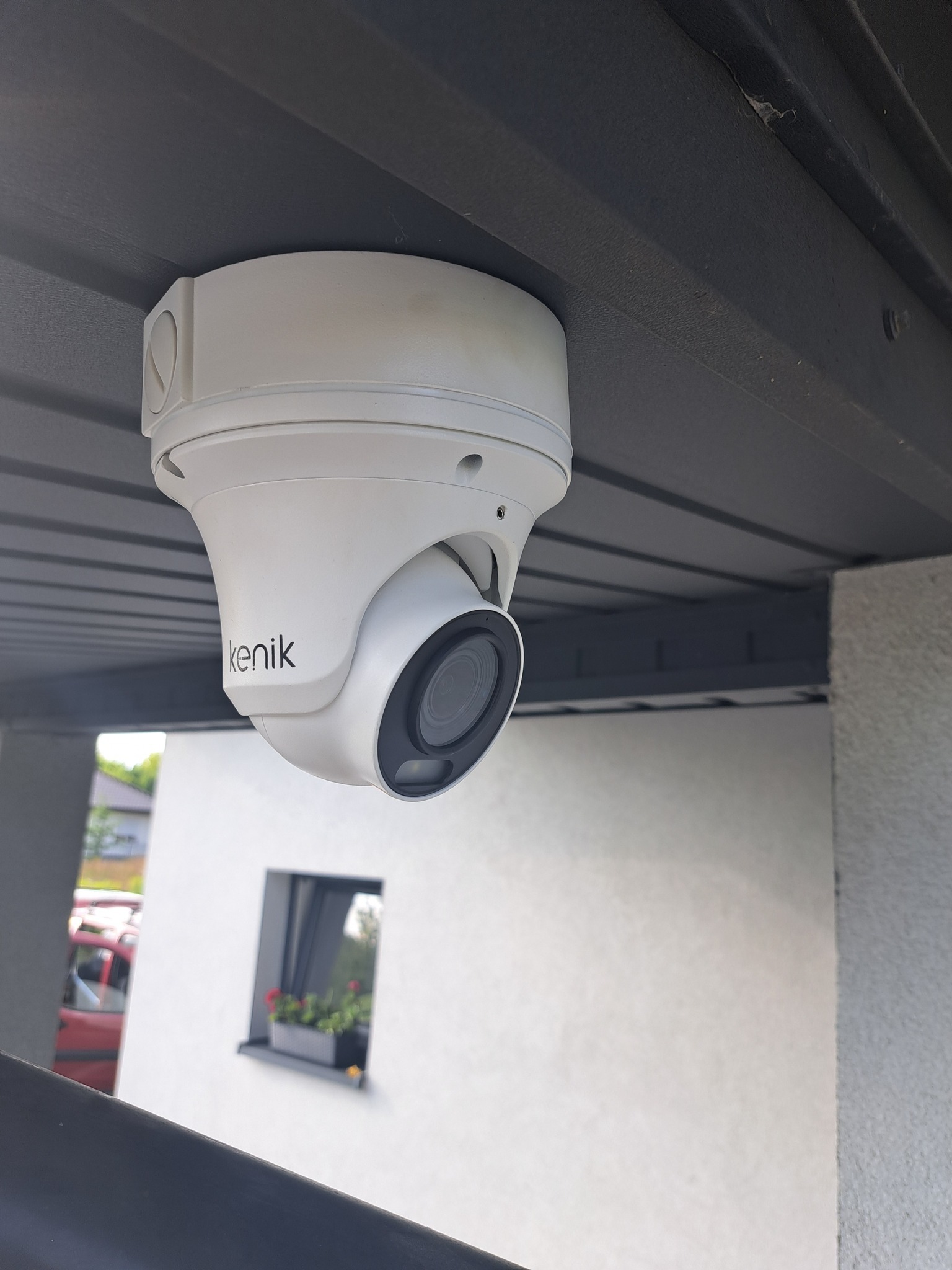 Kamera monitoringu Kenik, kopułkowa, zamontowana pod zadaszeniem. Widoczny fragment budynku z oknem i czerwony samochód w tle. Ujęcie z dołu.