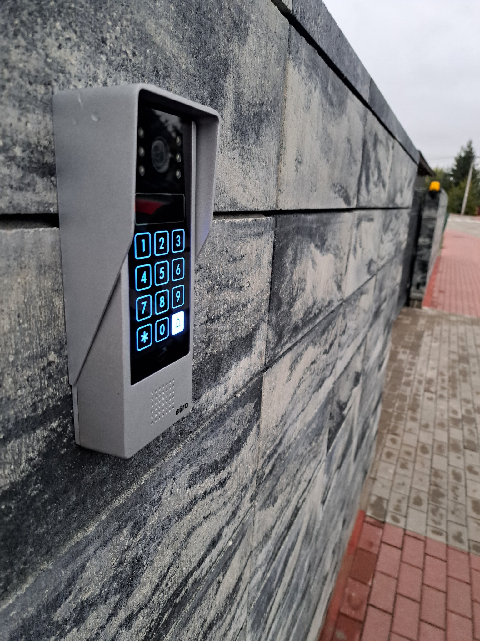 Szary wideodomofon Eura z podświetlaną klawiaturą numeryczną zamontowany na szarym, teksturowanym murze z kostki, widok z boku, oświetlenie dzienne.
