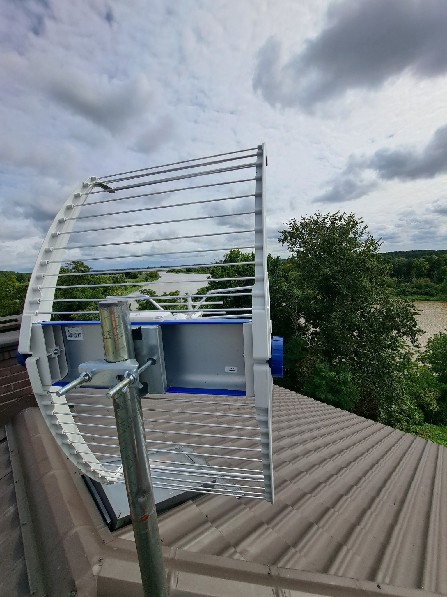 Antena kierunkowa zamontowana na dachu domu z widokiem na rzekę i zieleń. Metalowa konstrukcja anteny z elementami białymi i niebieskimi. Pochmurne niebo w tle.