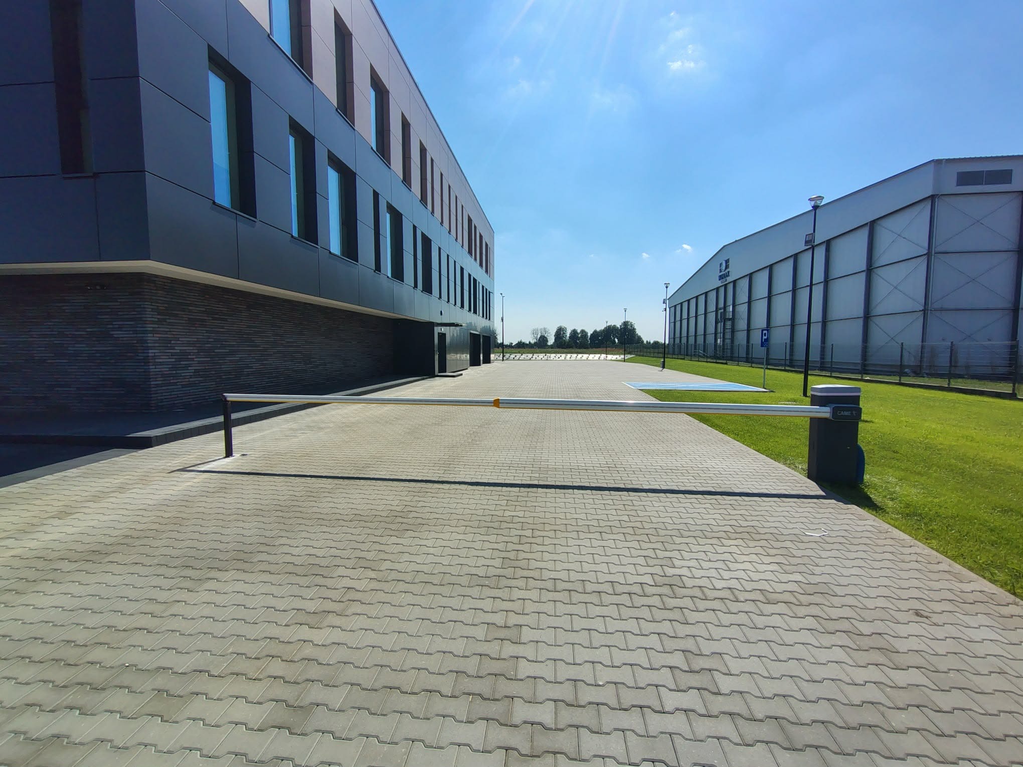 Zamknięty szlaban automatyczny przed nowoczesnym budynkiem biurowym i halą magazynową w słoneczny dzień. Kostka brukowa na pierwszym planie.