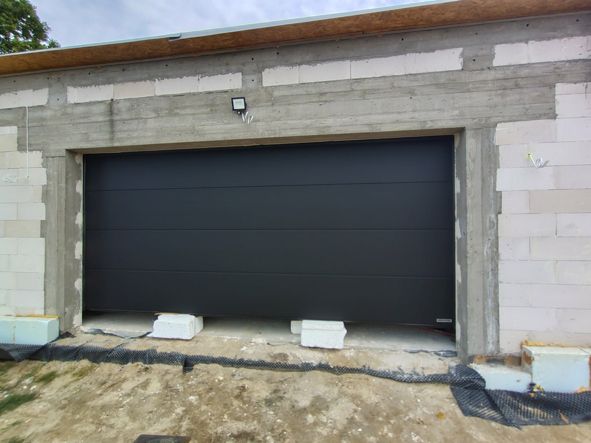 Zamknięta, segmentowa brama garażowa w surowym otoczeniu budowy, z widocznym oświetleniem zewnętrznym i blokami podtrzymującymi. Szara kolorystyka bramy.