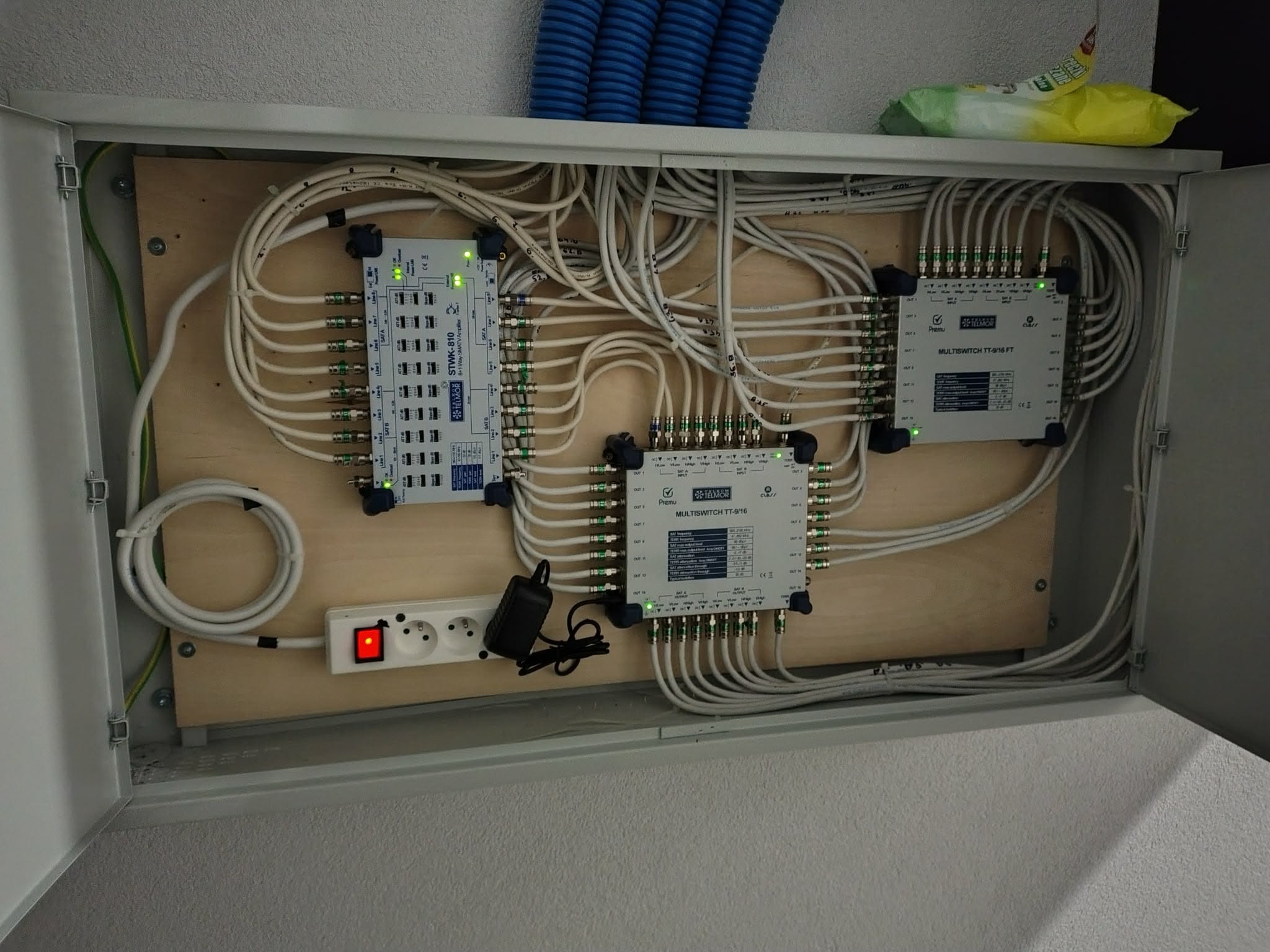 Rozdzielnica antenowa z multiswitchami i okablowaniem wewnątrz szafki. Widoczne złącza, kable koncentryczne i gniazdko elektryczne z włącznikiem.