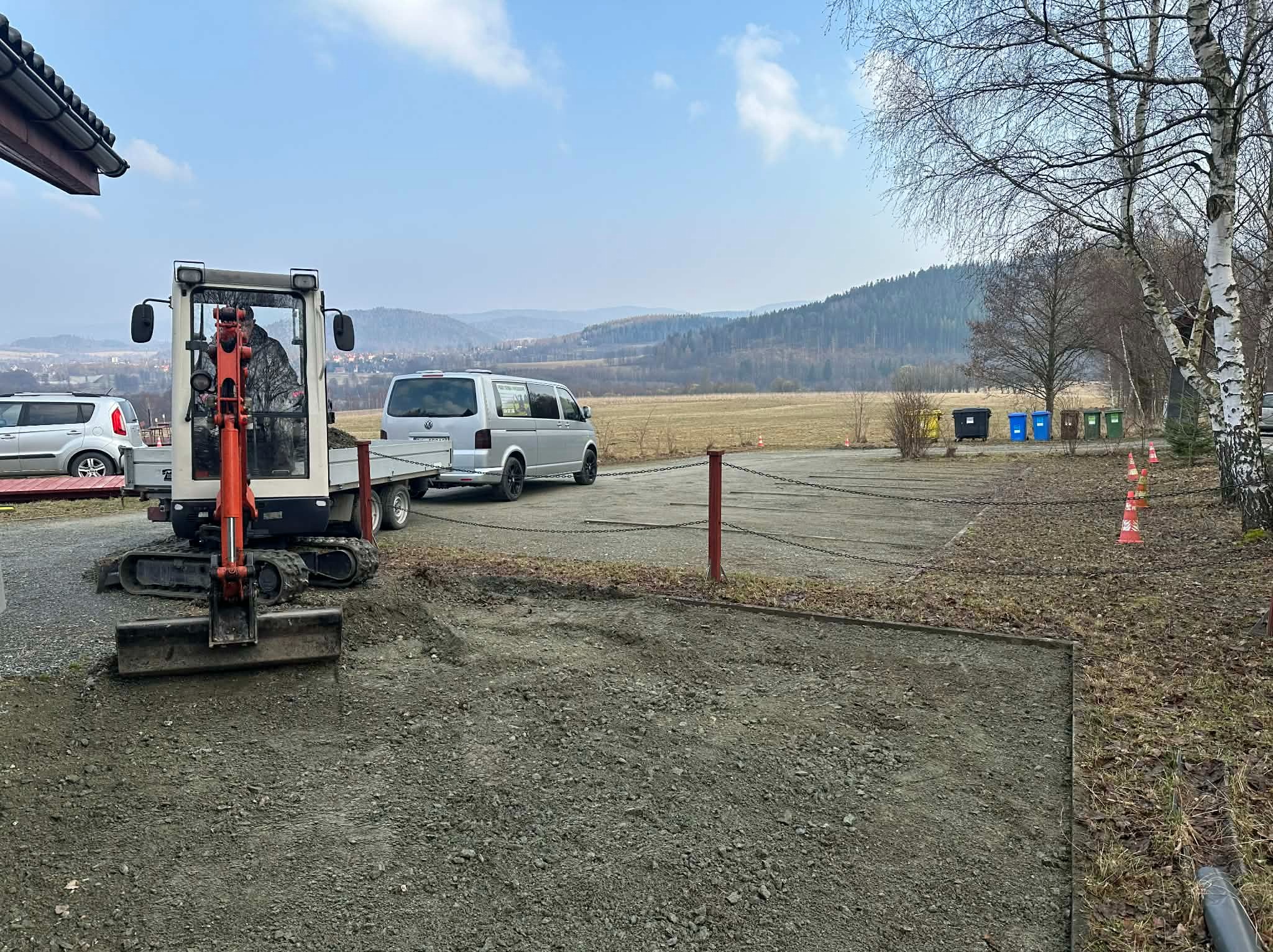 Mini koparka na przyczepie, przygotowuje teren pod budowę. W tle krajobraz zalesionych wzgórz i zaparkowany bus. Widoczne ślady pracy koparki na ziemi.