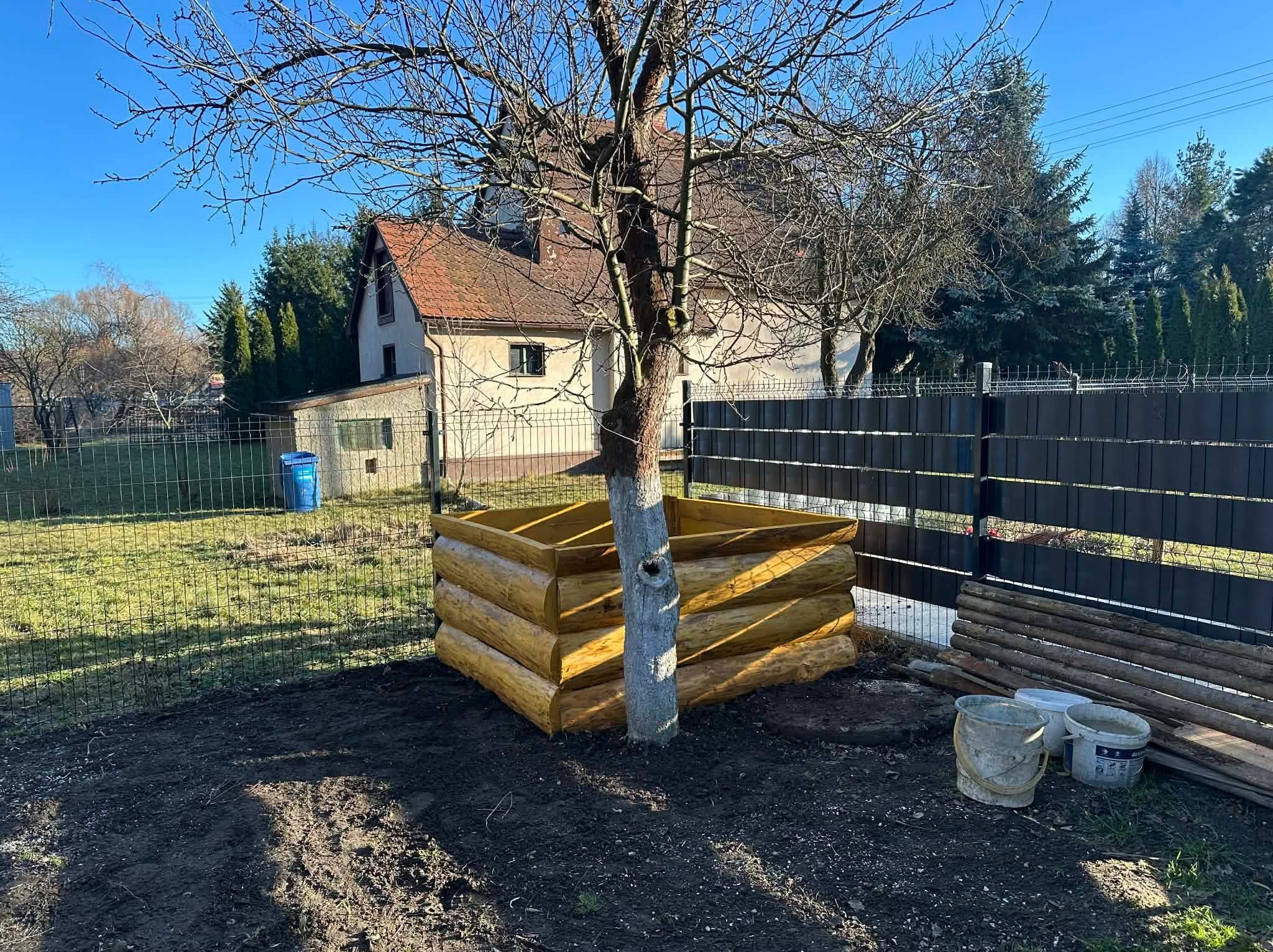 Obrzeże z drewnianych bali wokół drzewa na posesji, świeżo obsypane ziemią. W tle dom z czerwonym dachem i metalowe ogrodzenie.