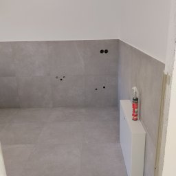 VIKI BUD - Szare płytki na ścianie i podłodze w łazience, widoczne otwory na armaturę i biała szafka pod umywalkę. Na szafce stoi puszka z pianką montażową.