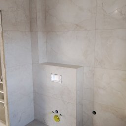 VIKI BUD - Wykończona łazienka z beżowymi płytkami imitującymi marmur. Widoczne podłączenia do toalety podtynkowej i otwór na rurę. Minimalistyczny styl.