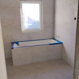 VIKI BUD - Jasna łazienka z wanną obłożoną płytkami imitującymi marmur, zabezpieczoną niebieską taśmą. Okno z widokiem na zieleń. Minimalistyczny design, świeżo po remoncie.