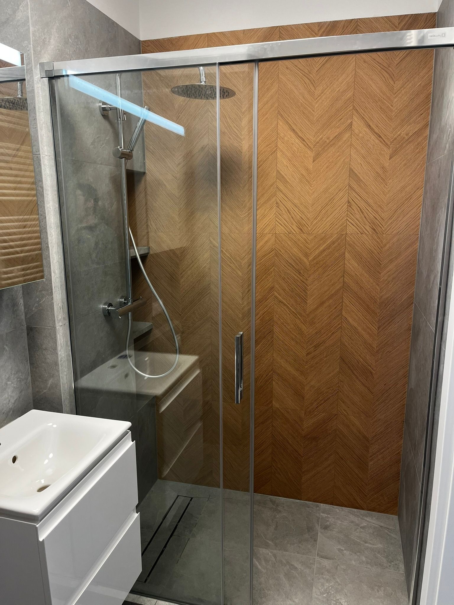 Nowoczesna kabina prysznicowa z przesuwnymi drzwiami, wykończona drewnopodobną mozaiką w jodełkę i szarą płytką imitującą kamień. Widoczna biała umywalka.