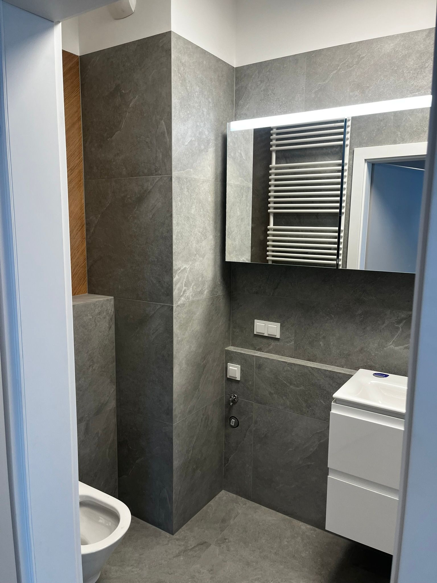 Nowoczesna łazienka w szarościach z podświetlanym lustrem, grzejnikiem drabinkowym i białą szafką z umywalką. Minimalistyczny design, płytki imitujące kamień.