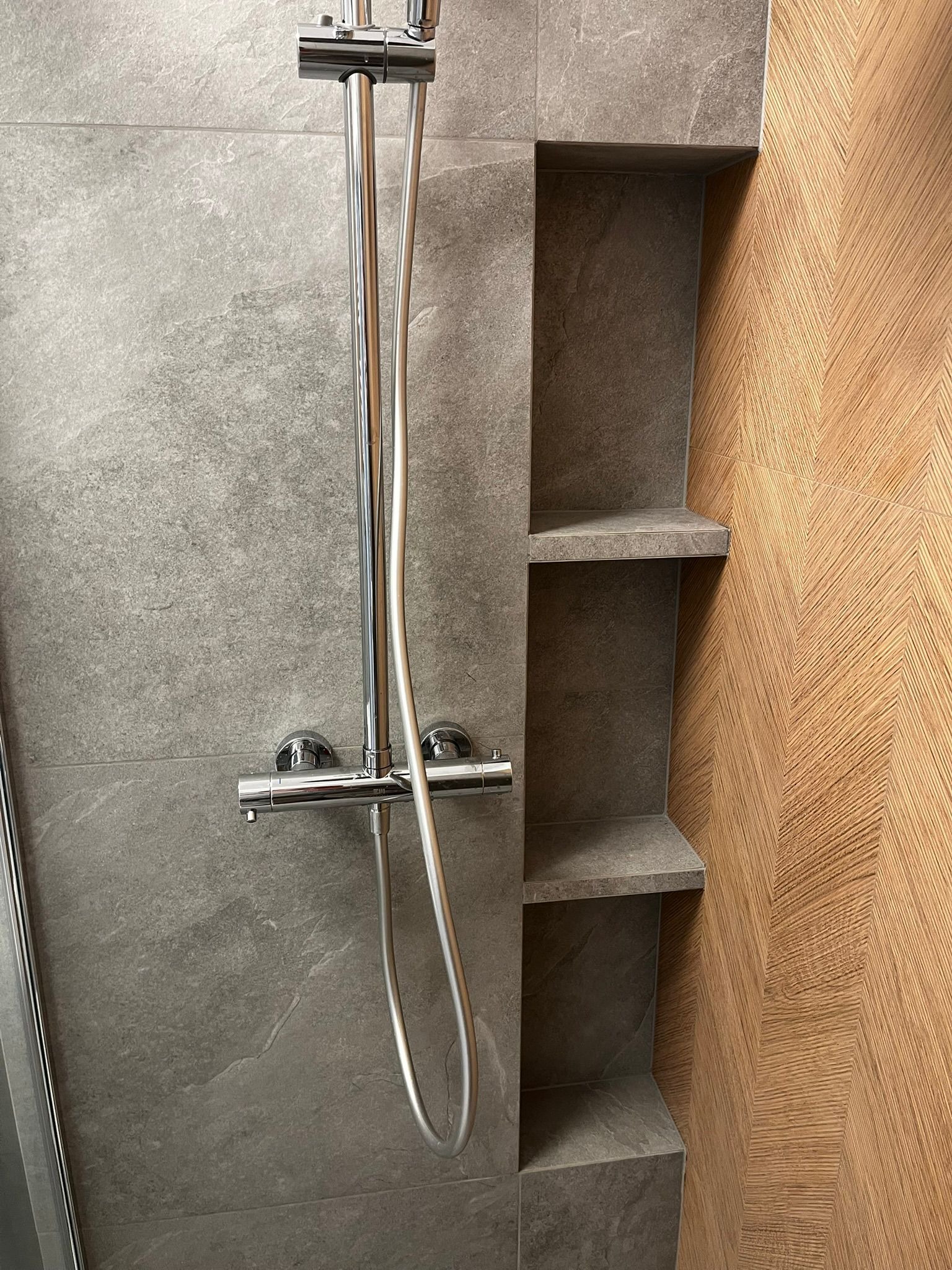 Nowoczesna łazienka z szarą, kamienną okładziną i drewnianą ścianą w jodełkę. Wnęka z półkami i chromowana bateria prysznicowa.