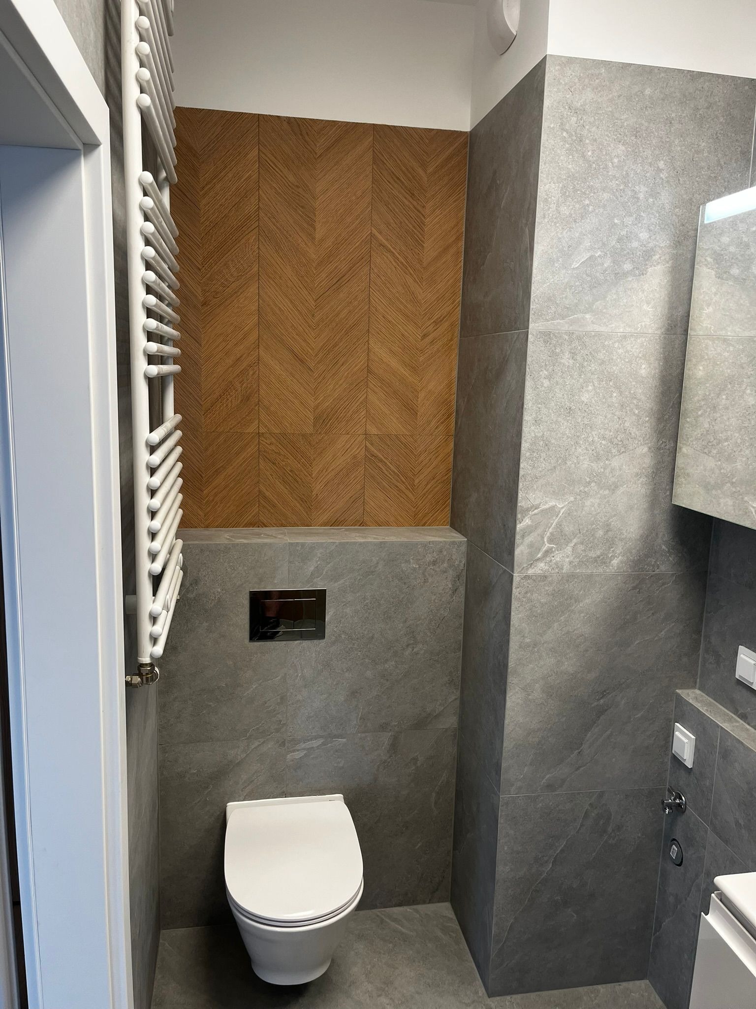 Nowoczesna łazienka z szarymi płytkami imitującymi kamień, drewnianą mozaiką na ścianie i białym grzejnikiem drabinkowym. Uwagę zwraca minimalistyczna toaleta podtynkowa.