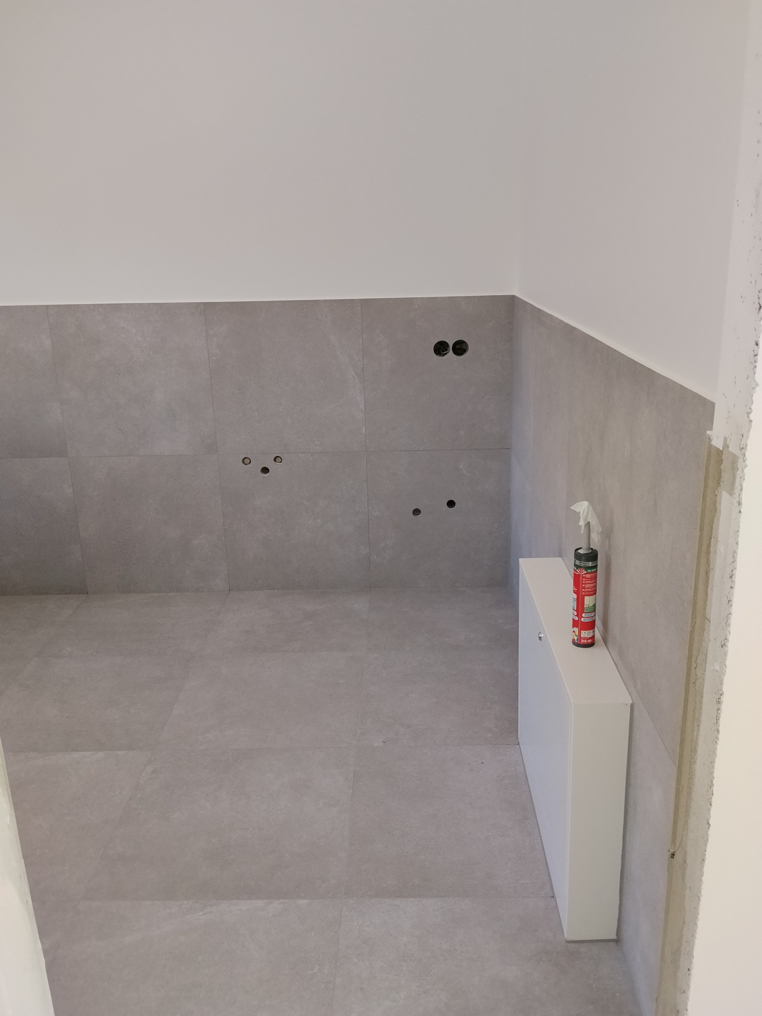 Szare płytki na ścianie i podłodze w łazience, widoczne otwory na armaturę i biała szafka pod umywalkę. Na szafce stoi puszka z pianką montażową.