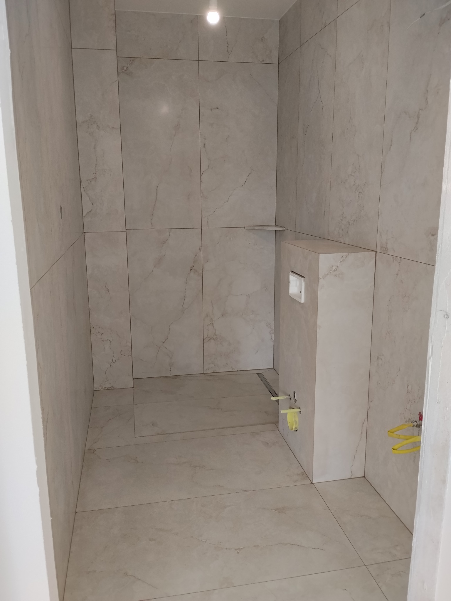 Wykończona łazienka z beżowymi płytkami imitującymi marmur, widoczny stelaż podtynkowy WC i odpływ liniowy. Minimalistyczny design, jasne oświetlenie.