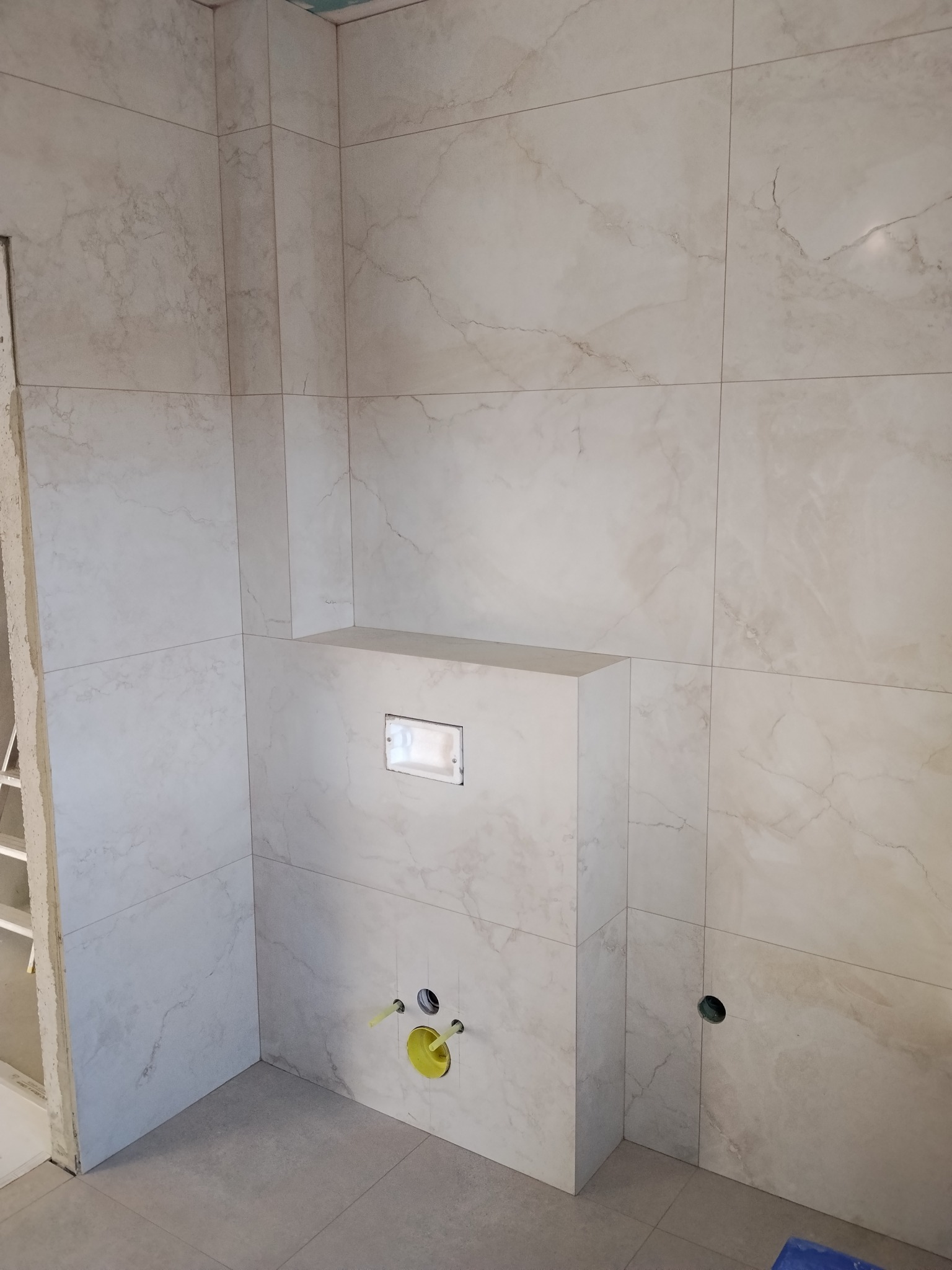 Wykończona łazienka z beżowymi płytkami imitującymi marmur. Widoczne podłączenia do toalety podtynkowej i otwór na rurę. Minimalistyczny styl.