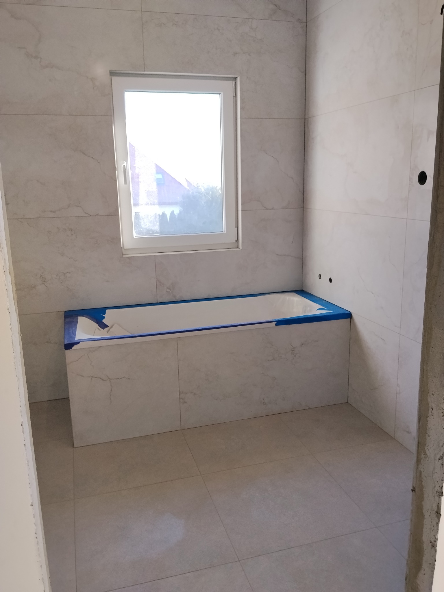 Jasna łazienka z wanną obłożoną płytkami imitującymi marmur, zabezpieczoną niebieską taśmą. Okno z widokiem na zieleń. Minimalistyczny design, świeżo po remoncie.