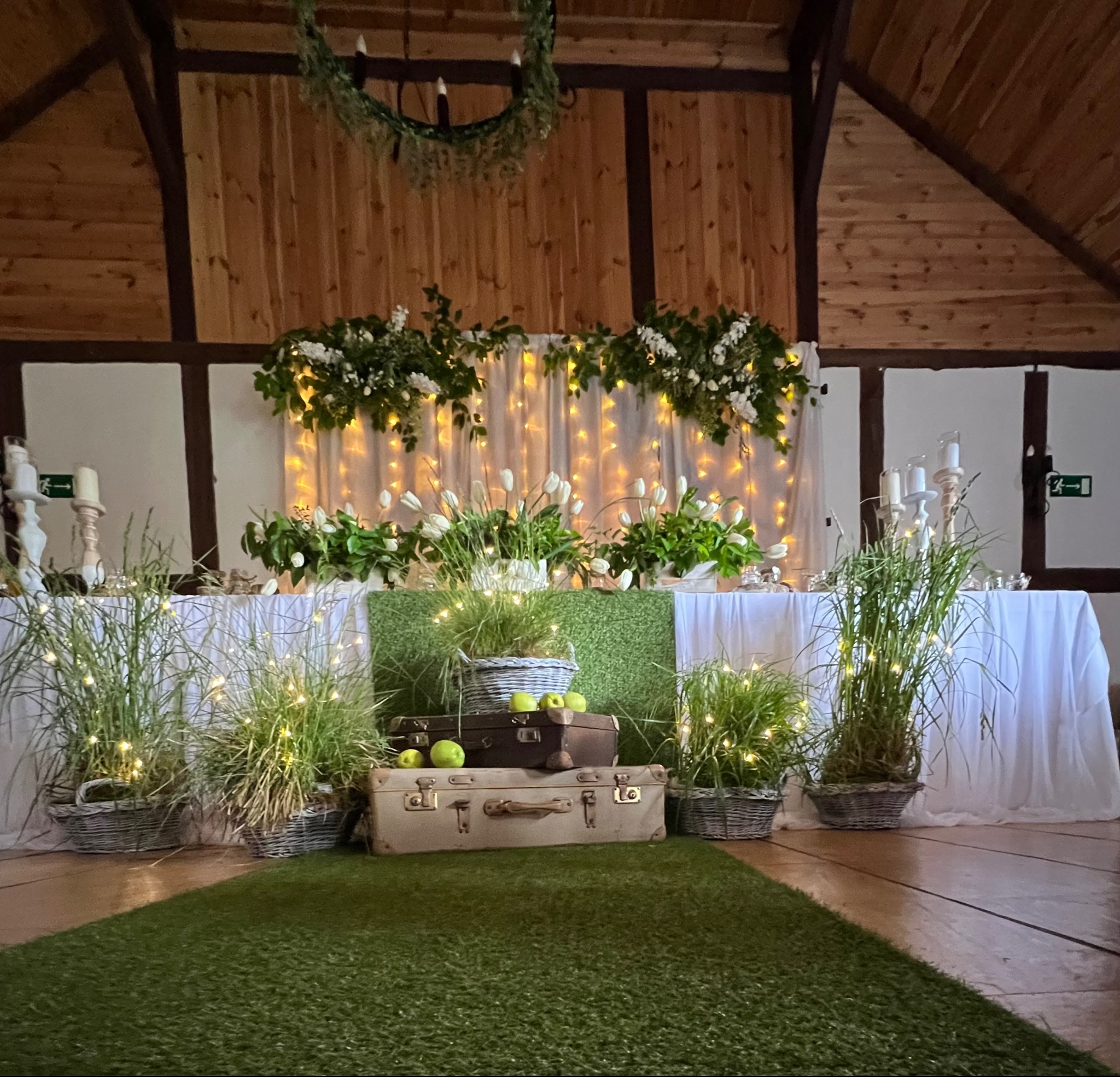 Dekoracja stołu weselnego w stylu rustykalnym: walizki vintage, trawy w koszach, jabłka i girlandy świetlne na tle drewnianej ściany.