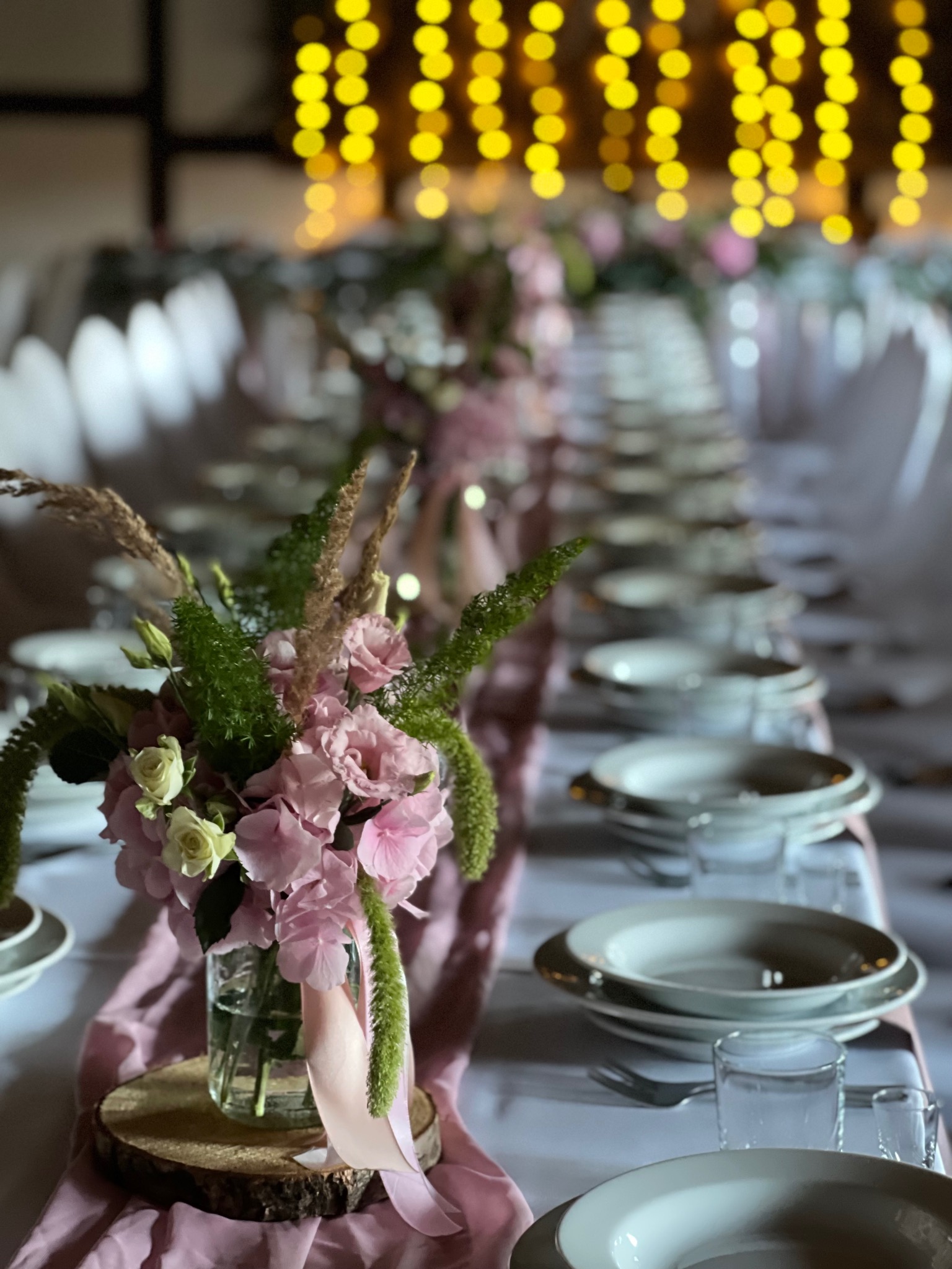 Udekorowany stół weselny z kwiatami w szklanym wazonie na drewnianym plastrze, różowy bieżnik, zastawa stołowa i ciepłe światła w tle. Dekoracja stołu.