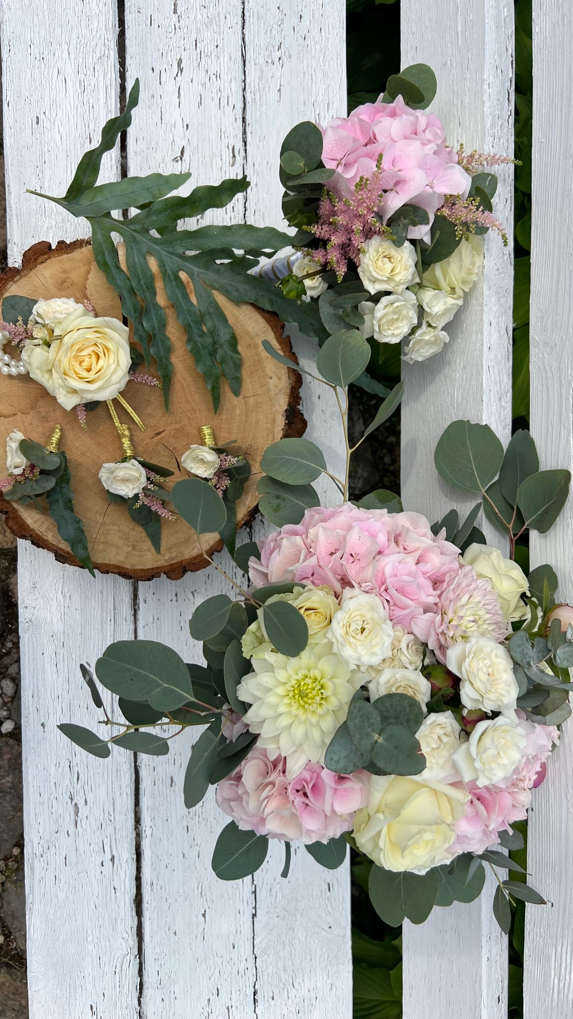 Kompozycja kwiatowa z róż, hortensji i eukaliptusa na białym, drewnianym tle. Bukiet ślubny i butonierki na plastrze drewna. Delikatne, pastelowe barwy.