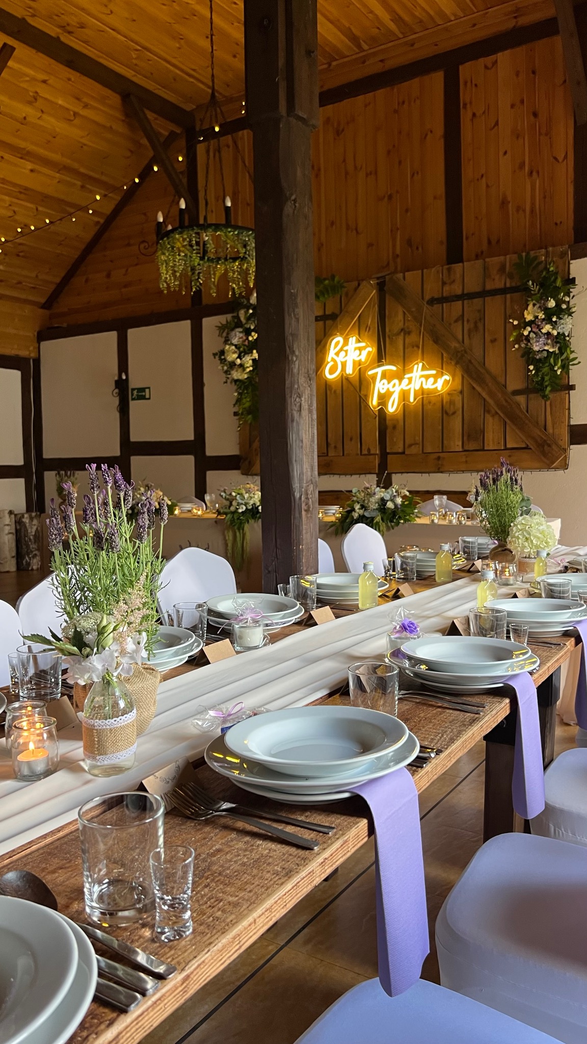 Uroczysta aranżacja stołu weselnego w rustykalnej sali: drewniany stół, biała zastawa, fioletowe akcenty, kwiaty i neon 'Better Together' na drewnianych drzwiach.