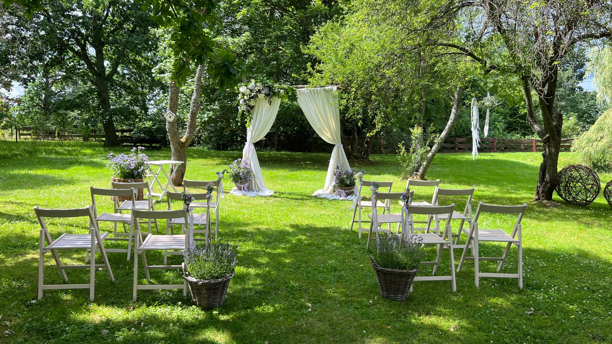 Uroczysta aranżacja ślubna w plenerze: biały baldachim z kwiatami, krzesła dla gości, dekoracje z lawendy i lampiony na drzewach w słoneczny dzień.