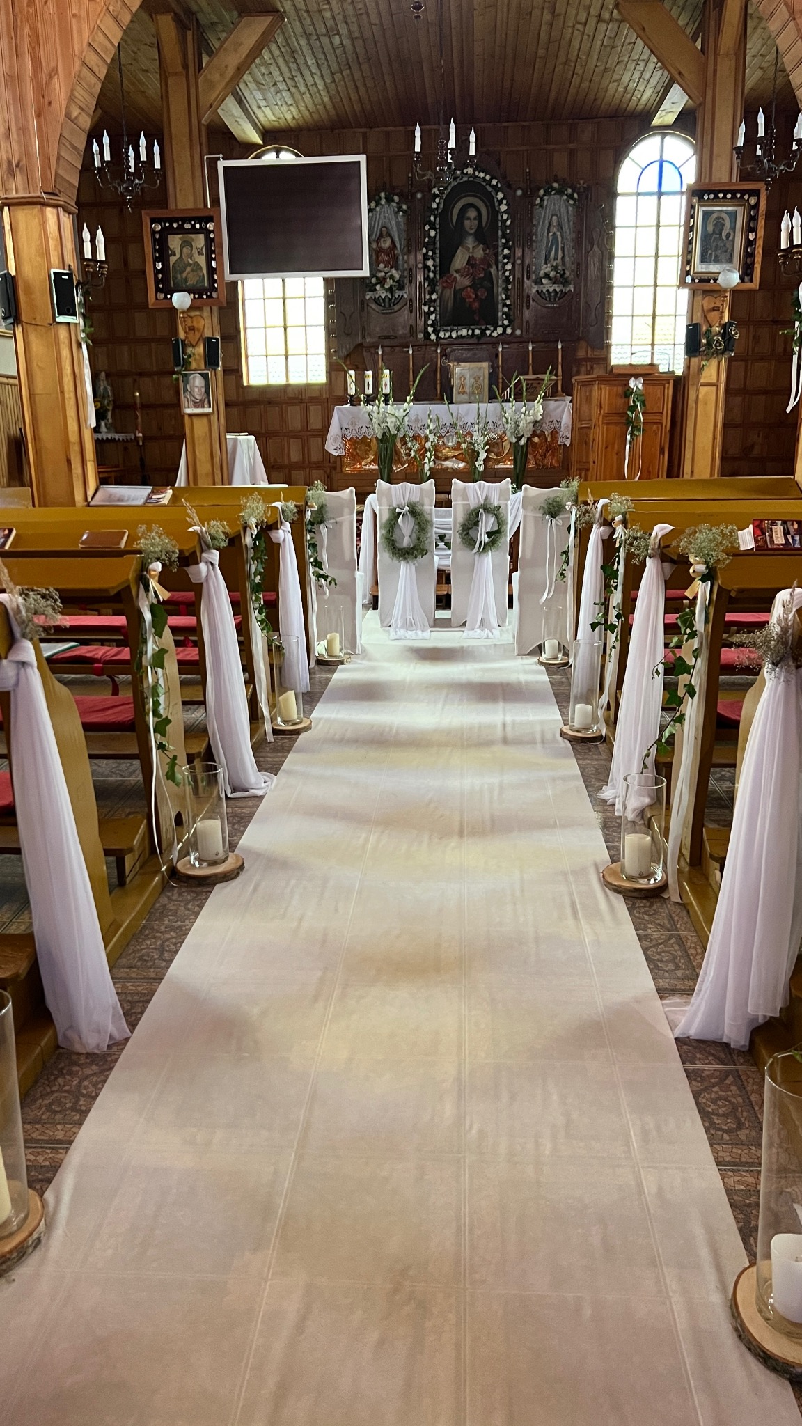 Udekorowany kościół na ślub: biały dywan, wstążki, świece i wianki na krzesłach. Drewniane wnętrze kościoła z obrazami i oświetleniem.