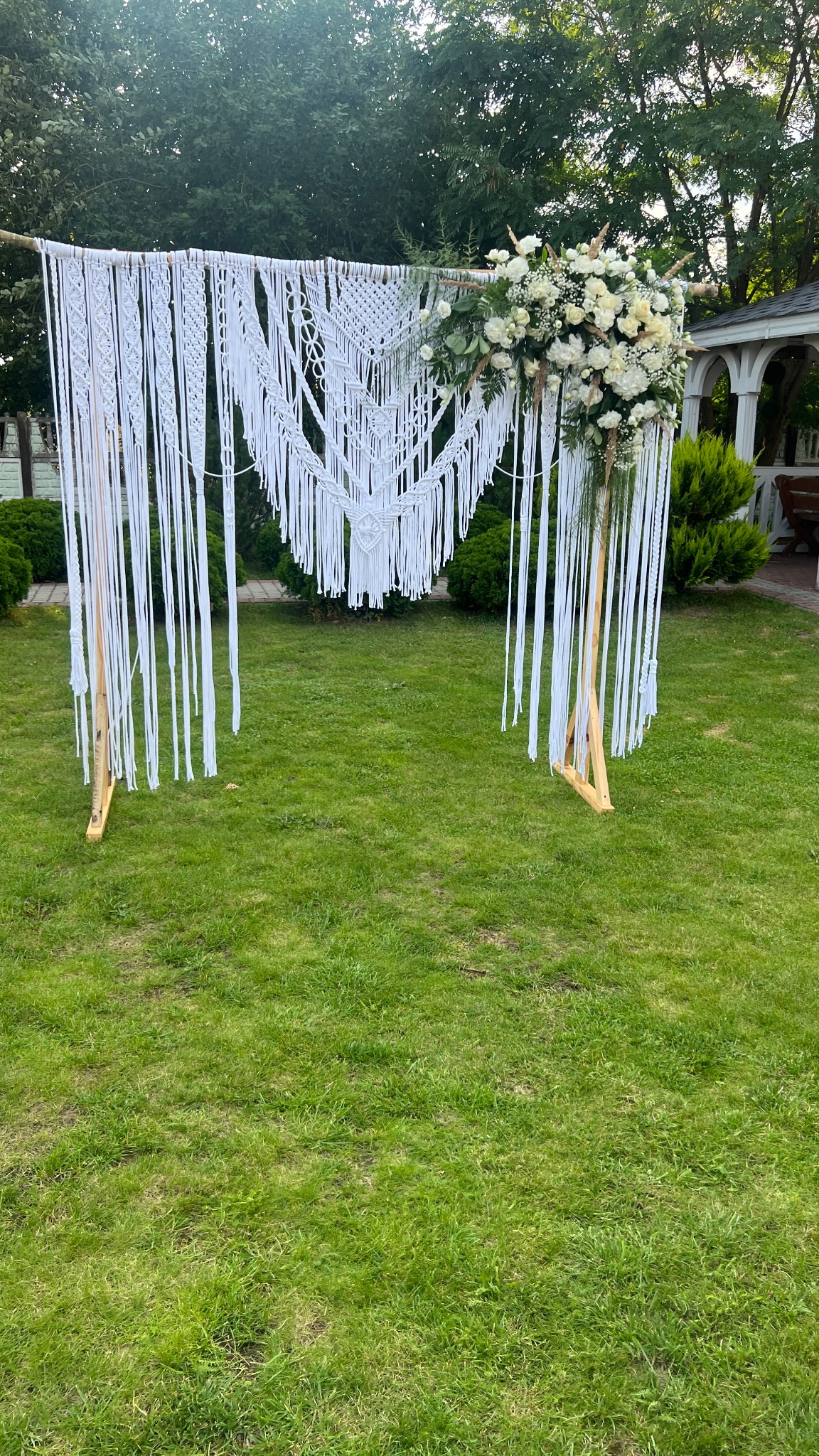 Dekoracja ślubna w stylu boho: białe makramy i kompozycja z białych kwiatów na drewnianej konstrukcji, ustawiona na trawniku przed altaną.