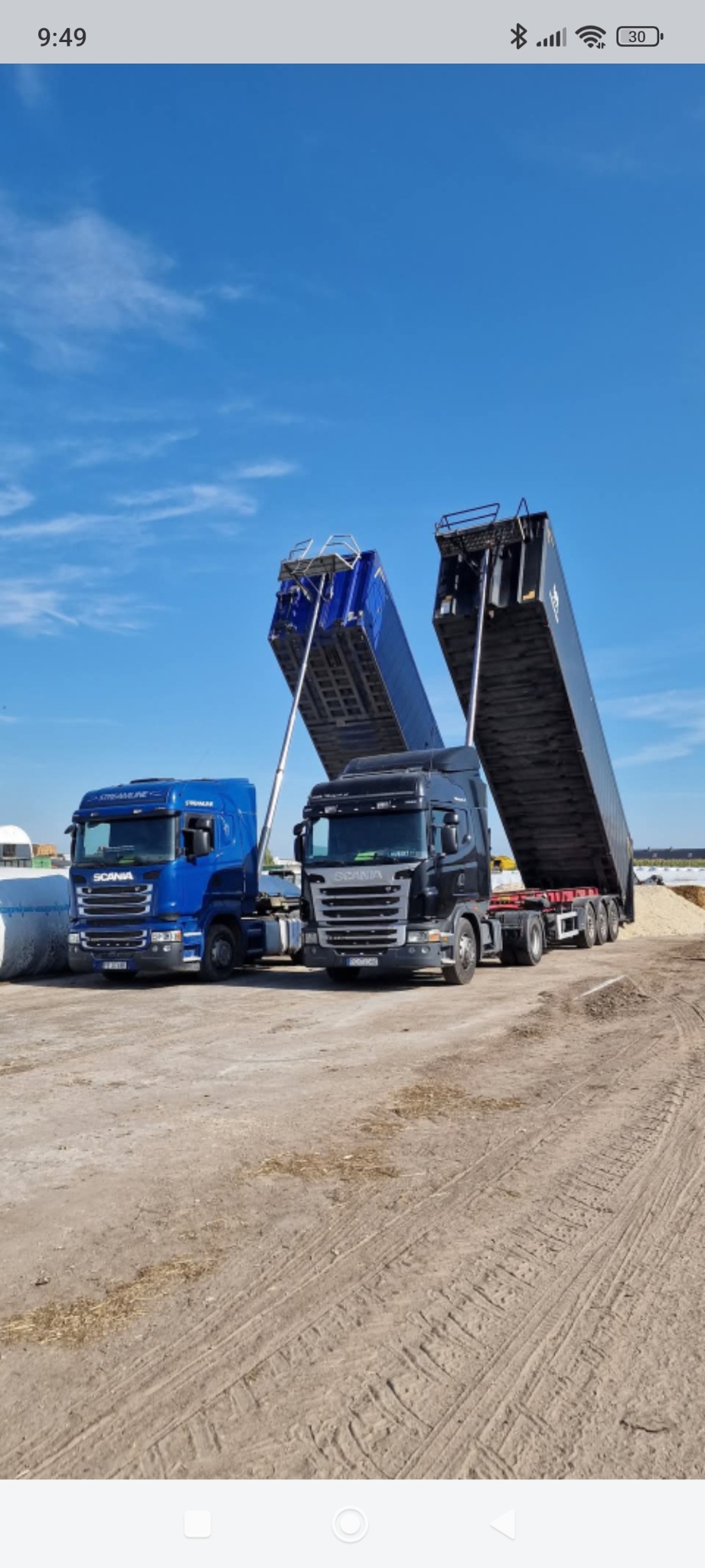 Dwa samochody ciężarowe Scania z otwartymi naczepami, jeden niebieski, drugi czarny, na placu pod błękitnym niebem. Widoczne ślady opon na ziemi.