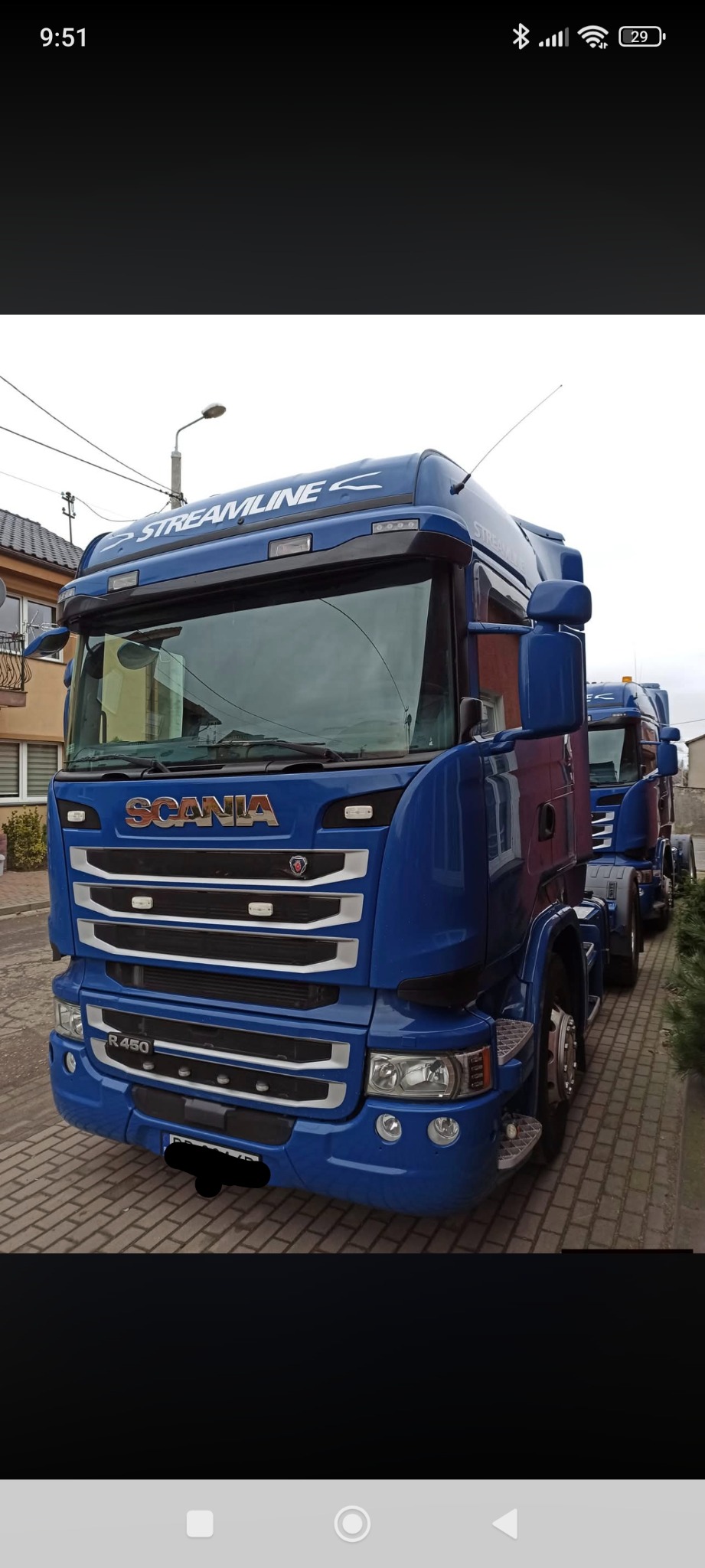 Błękitny ciągnik siodłowy Scania R450 Streamline na tle budynku. Widoczne detale: chromowane elementy, antena, tablica rejestracyjna (częściowo zasłonięta).