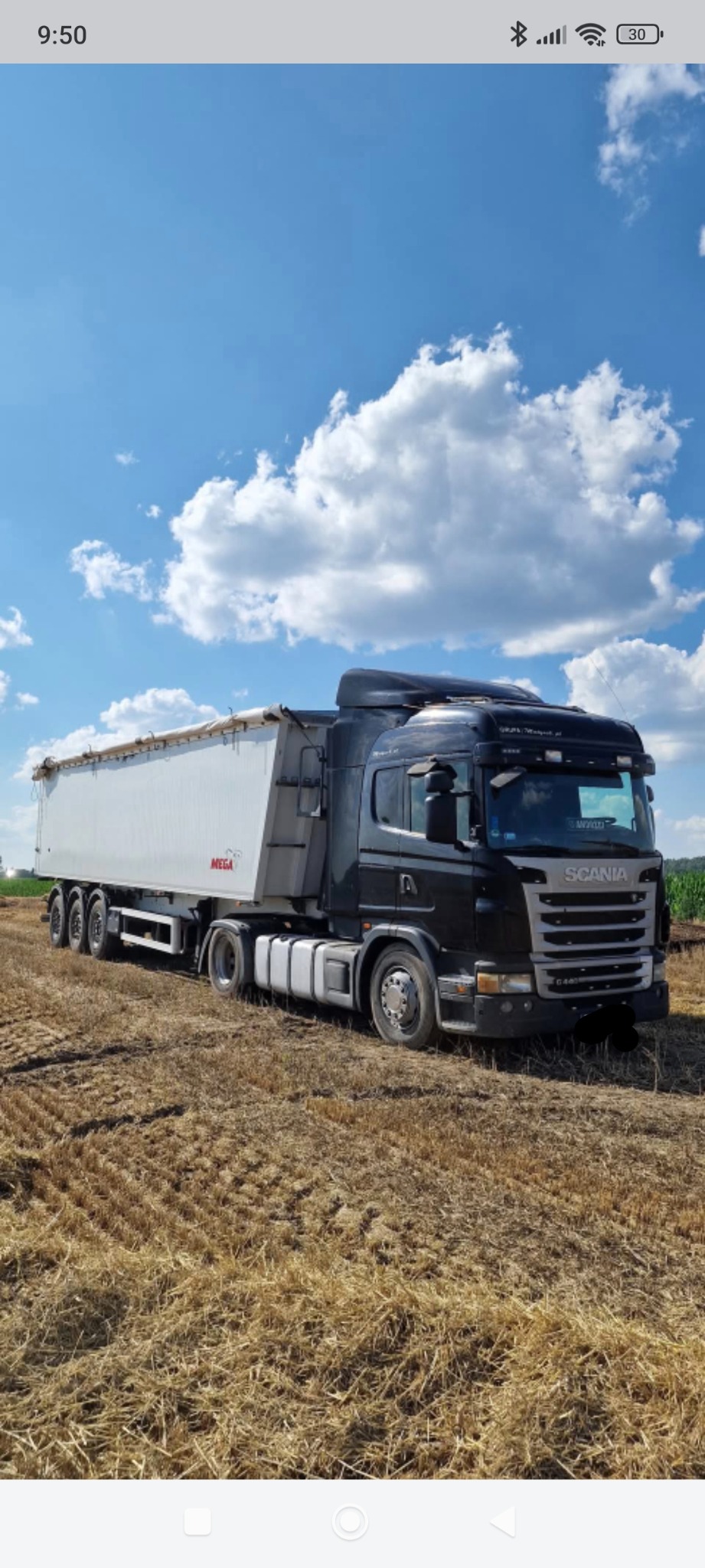 Czarna ciężarówka Scania z białą naczepą MEGA na polu ścierniska pod błękitnym niebem z chmurami. Widok z boku, lekko z dołu.