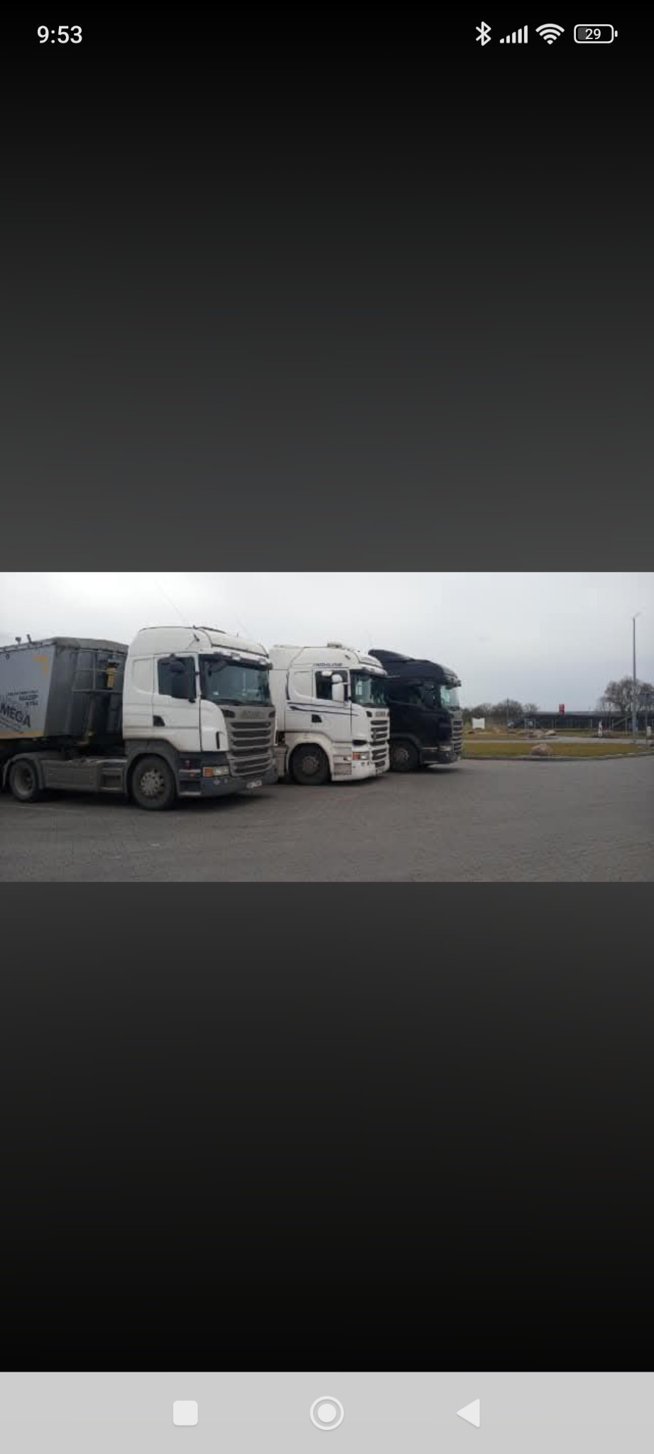 Trzy ciężarówki Scania różnych kolorów (biała, biała z czarnymi elementami i czarna) zaparkowane na parkingu. Widok z boku, pochmurne niebo w tle.